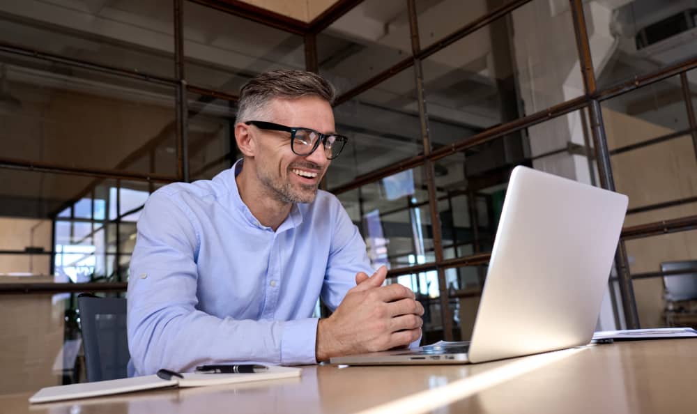 Mann mit Brille und blauen Hemd sitzt am Schreibtisch vor seinem Laptop