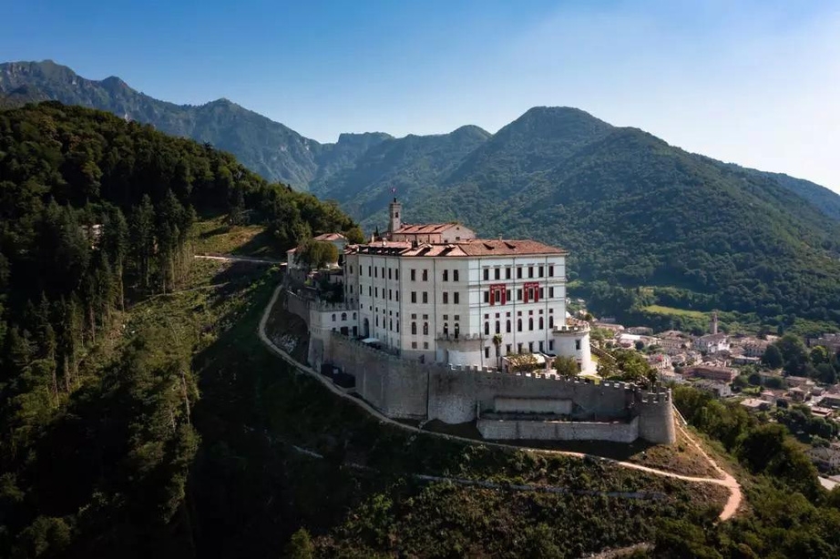 Eine große alte Festung thront auf einem Hügel, umgeben von üppigen grünen Bergen. Das Bauwerk überblickt eine kleine Stadt im Tal darunter. Über ihnen ist ein klarer, blauer Himmel zu sehen.