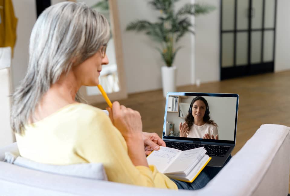 Frau mit Notizbuch und Stift sitzt vor ihrem Laptop und spricht in einer Videokonferenz mit einer weiteren Frau