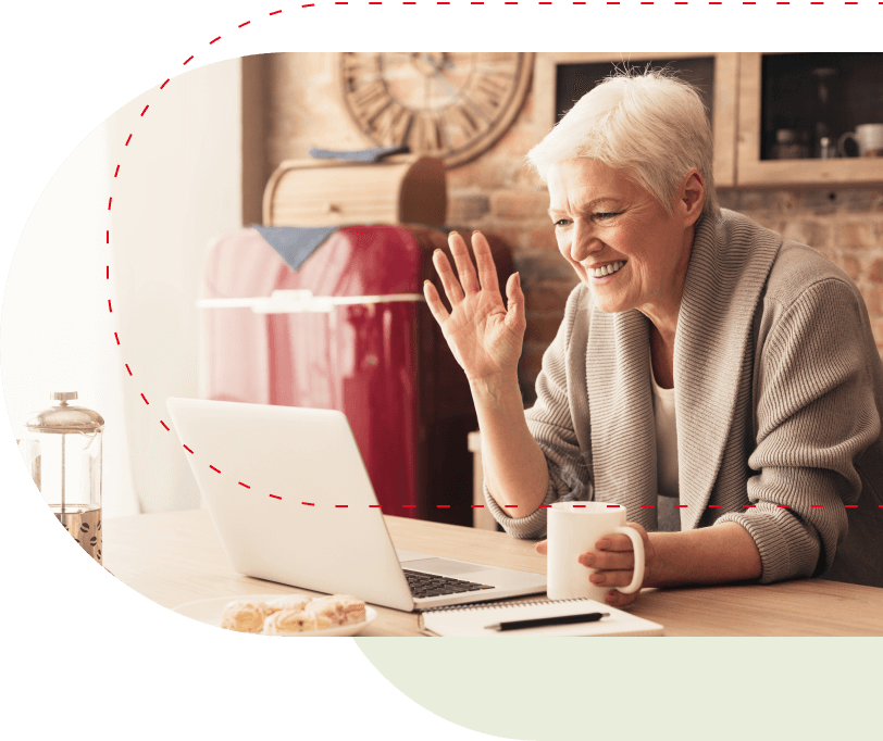 Ältere Dame lernt online Italienisch auf A1-Niveau als Anfängerin. Sie sitzt vor dem Laptop und hebt die Hand zur Begrüßung für Ihre erste Italienisch Stunde. In der anderen Hand hält sie eine Tasse mit Kaffee.