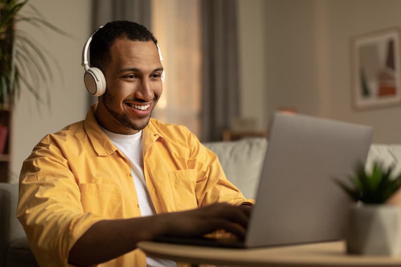 man mit gelbem Hemd und weißem Kopfhörer sitzt vorm Computer, lächelt und lernt italienisch