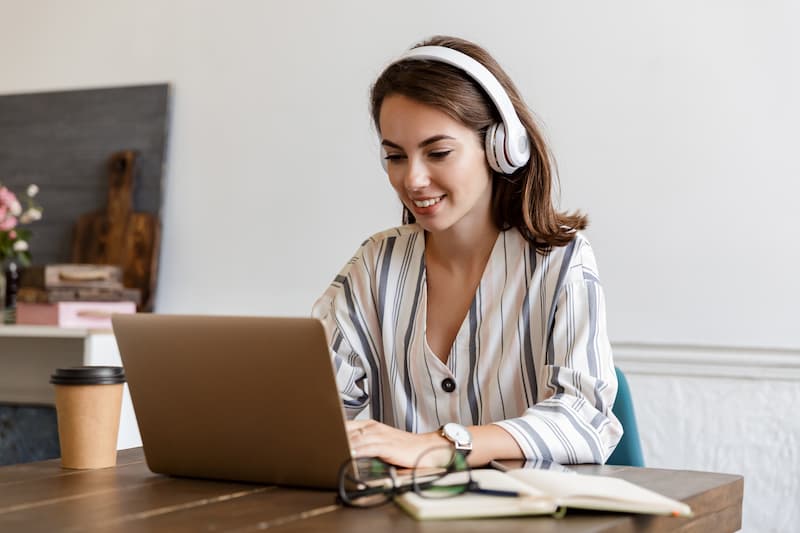 Frau lächelt und sitzt am Laptop mit weißen Kopfhörer und lernt italienisch