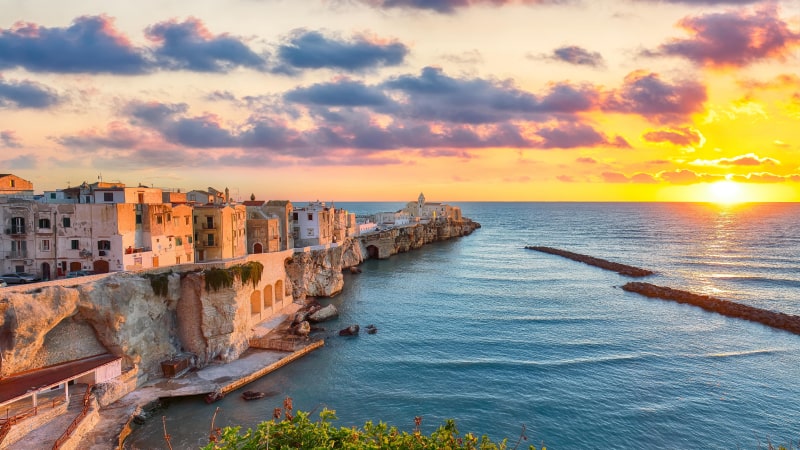 Lebendiger Sonnenuntergang über der Küstenstadt Vieste mit historischen Gebäuden an Klippenrändern, ruhigem blauen Wasser und vereinzelten Wolken am Himmel. Grüne Sträucher im Vordergrund.