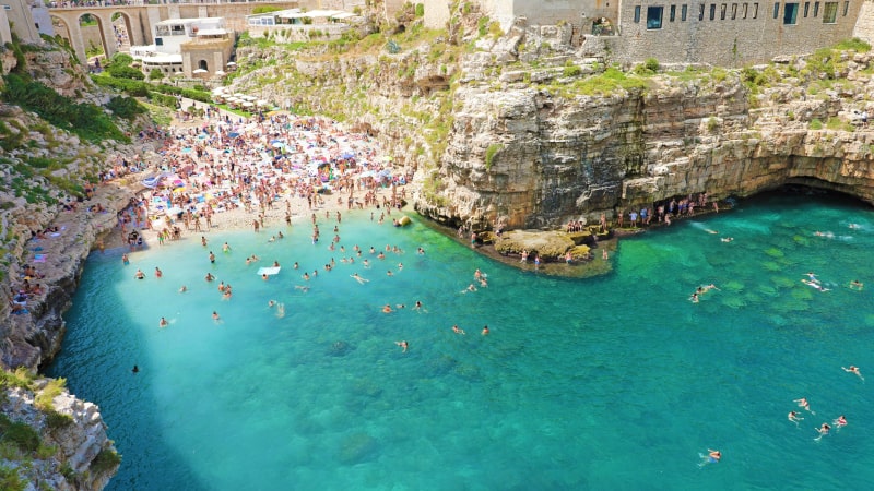 Ein überfüllter Strand mit türkisfarbenem Wasser und Klippen in Polignano a Mare, Italien. Menschen schwimmen im Wasser und sonnen sich am Ufer mit bunten Sonnenschirmen und Gebäuden im Hintergrund.