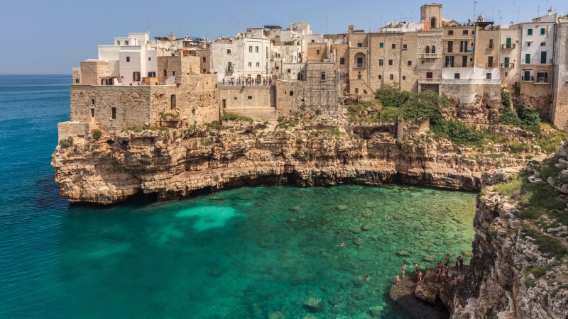 Die Küstenstadt Bari mit Steingebäuden liegt auf einer Felsklippe mit Blick auf das klare, blaue Meer. Unten schwimmen Menschen, und einige stehen auf den Felsen.