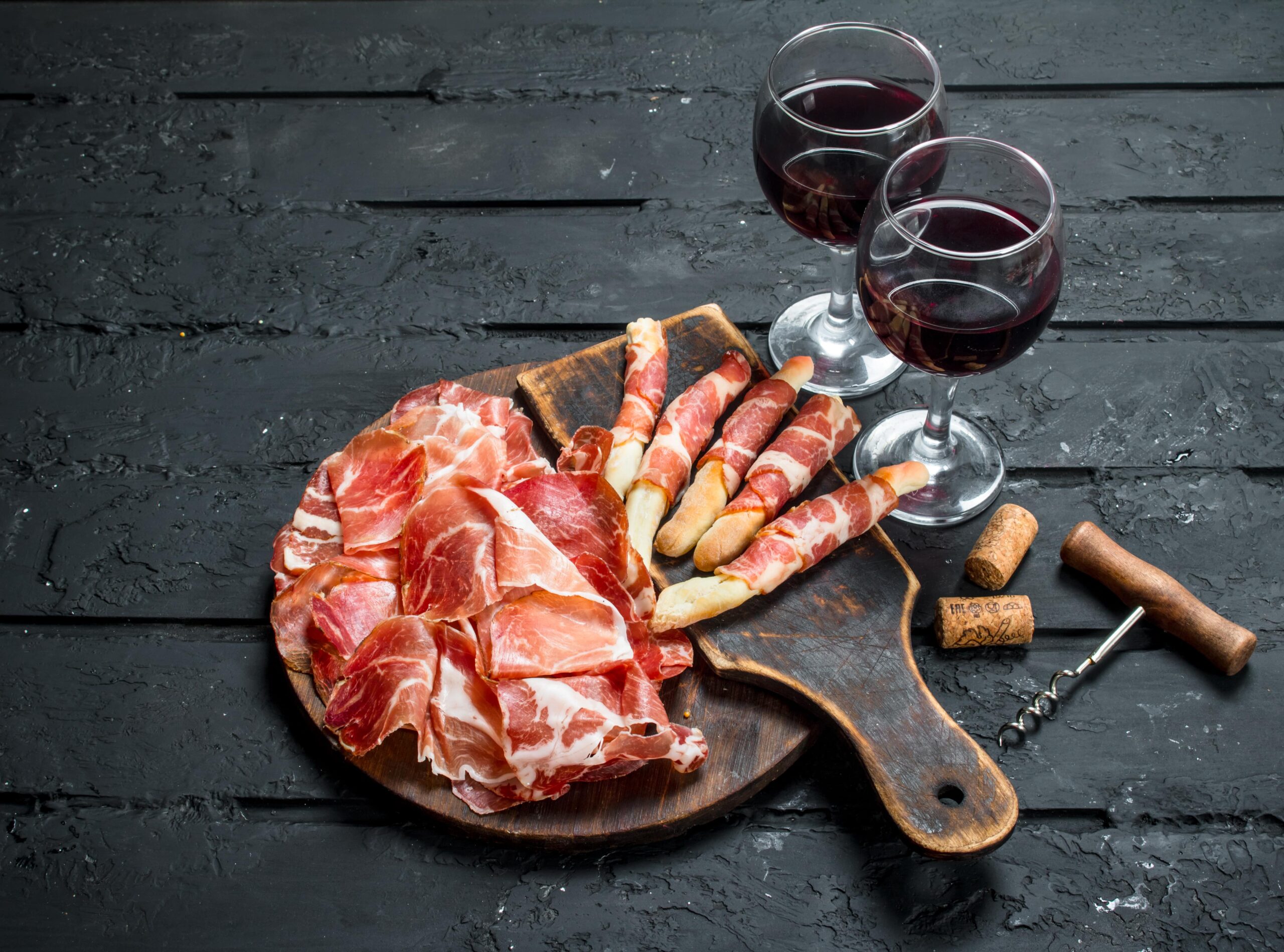 Ein Holzbrett mit Wurstscheiben und mit Wurst umwickelten Grissini, dazu zwei Gläser Rotwein, ein Korkenzieher und zwei Weinkorken auf dunklem Untergrund.