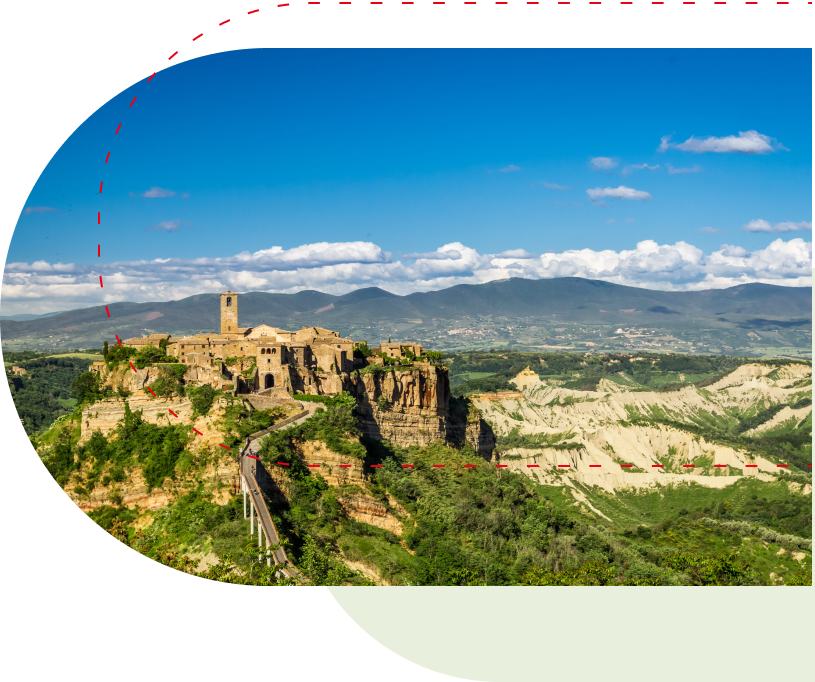 Auf einem Berggipfel gelegene Stadt mit Steingebäuden und einem Turm, umgeben von üppigem Grün und in der Ferne Bergen unter einem klaren blauen Himmel.