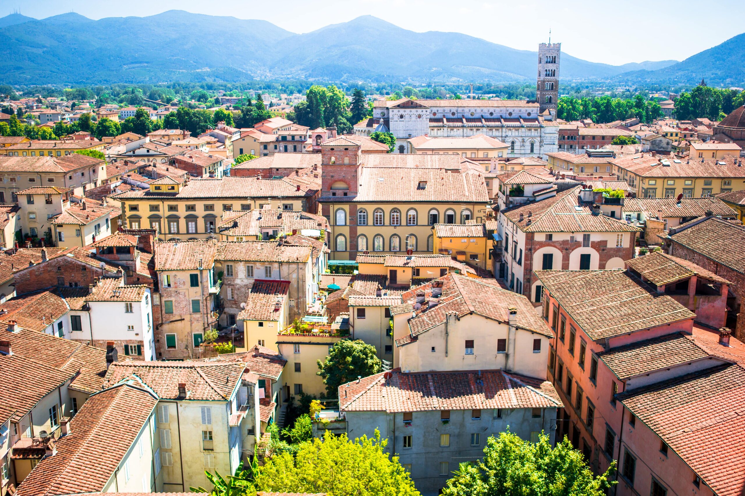 Luftaufnahme der historischen Stadt Lucca mit Terrakotta-Dächern, engen Gassen und einem zentralen Glockenturm. Im Hintergrund sind Hügel und Grünflächen zu sehen.