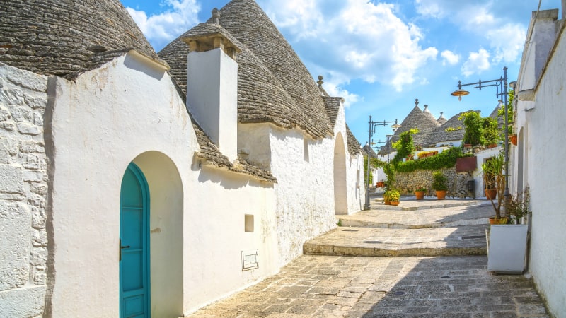 Schmale Kopfsteinpflasterstraße in Alberobello gesäumt von weißen Steinhäusern mit kegelförmigen Dächern, einer blauen Tür und Topfpflanzen unter einem blauen Himmel mit vereinzelten Wolken.