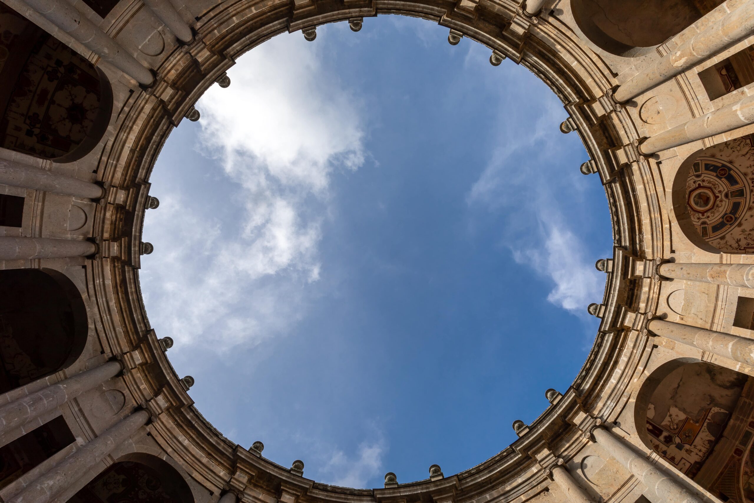 Blick auf einen blauen Himmel durch eine kreisförmige Öffnung in einer historischen Kolonnade mit detaillierter Steinarchitektur.