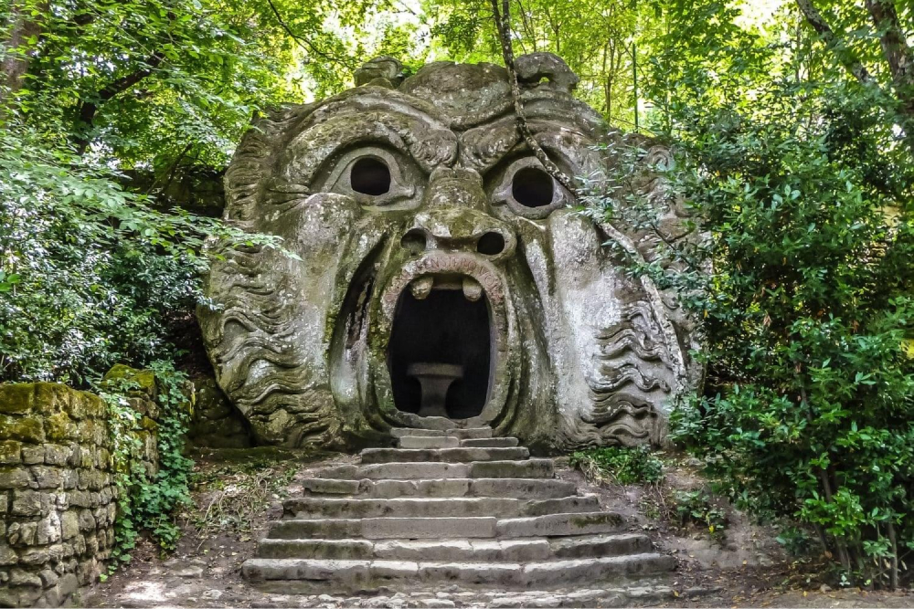 Eine Steinskulptur eines monströsen Gesichts mit offenem Mund, die am oberen Ende einer Treppe einen Eingang bildet, umgeben von üppigem Grün.