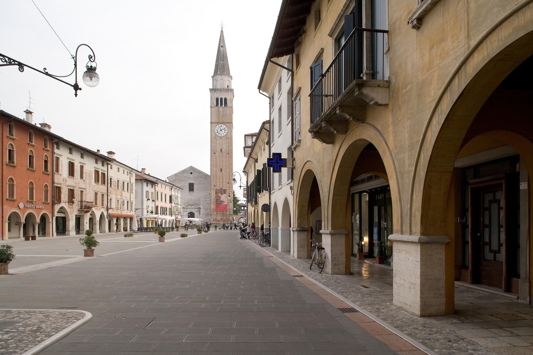 Eine ruhige Straßenszene in einer europäischen Stadt mit einem hohen Uhrenturm im Hintergrund, umgeben von mehrstöckigen Gebäuden mit gewölbten Gehwegen und ein paar am Bürgersteig geparkten Fahrrädern.