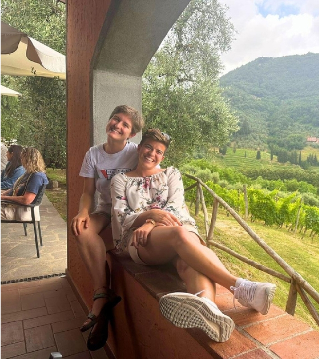 Zwei Menschen sitzen lächelnd auf einem Verandageländer, mit einer malerischen Aussicht auf üppig grüne Weinberge und sanfte Hügel im Hintergrund.