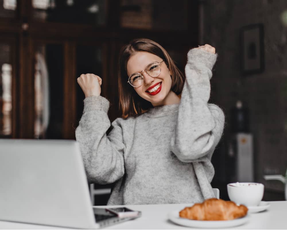 Frau mit Brille lacht, während sie vor einem Laptop sitzt mit einem Croissant und Kaffee