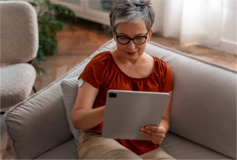 Person mit Kurzhaarfrisur und Brille sitzt entspannt auf grauem Sofa und nutzt ein Tablet.