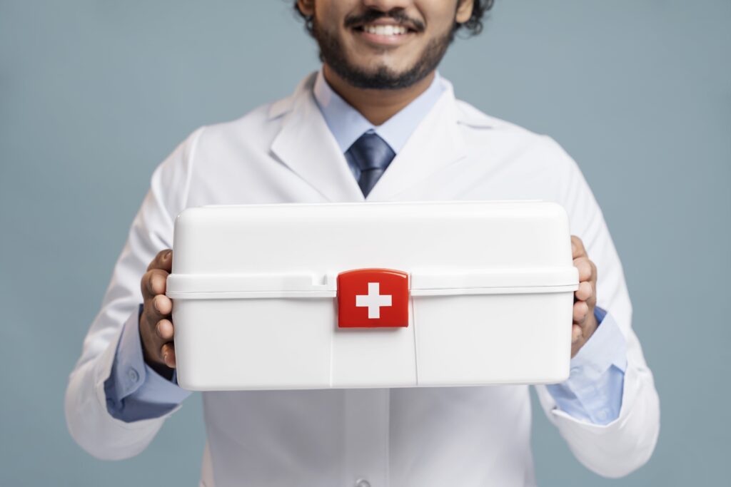 Person im Laborkittel, die vor einem einfarbigen Hintergrund ein weißes Erste-Hilfe-Set mit einem roten Kreuz hält.