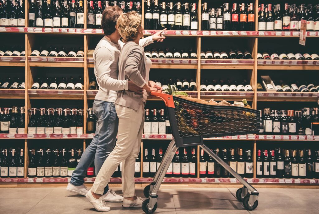 Ein Paar in Freizeitkleidung steht in einem Supermarktgang und wählt Wein aus Regalen voller verschiedener Flaschen aus. Vor ihnen steht ein Einkaufswagen.