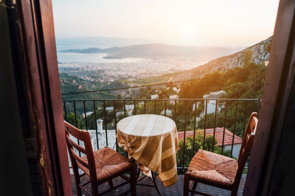 Ein kleiner runder Tisch mit karierter Tischdecke und zwei Holzstühle auf einem Balkon blicken auf eine hügelige Landschaft und eine entfernte Küste unter einem hellen Himmel.