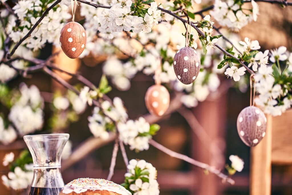Gepunktete Ostereier hängen an einem blühenden Ast; im Vordergrund sind ein Glaskrug und ein dekorierter Kuchen zu sehen, die die fröhliche Osterstimmung in Italien einfangen.