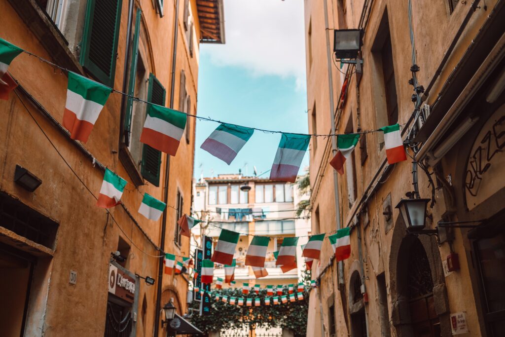 Schmale Straße in Italien, gesäumt von Gebäuden und zahlreichen italienischen Flaggen, die an Schnüren über ihnen hängen und lokale Bräuche und Traditionen feiern, während darüber ein klarer blauer Himmel sichtbar ist.