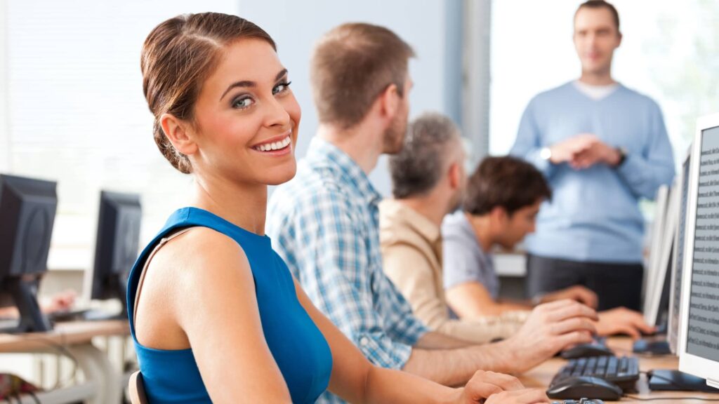 Eine Frau sitzt im Büro am Schreibtisch und lächelt. Die Umgebung ist formell und andere Personen arbeiten und unterhalten sich im Hintergrund.