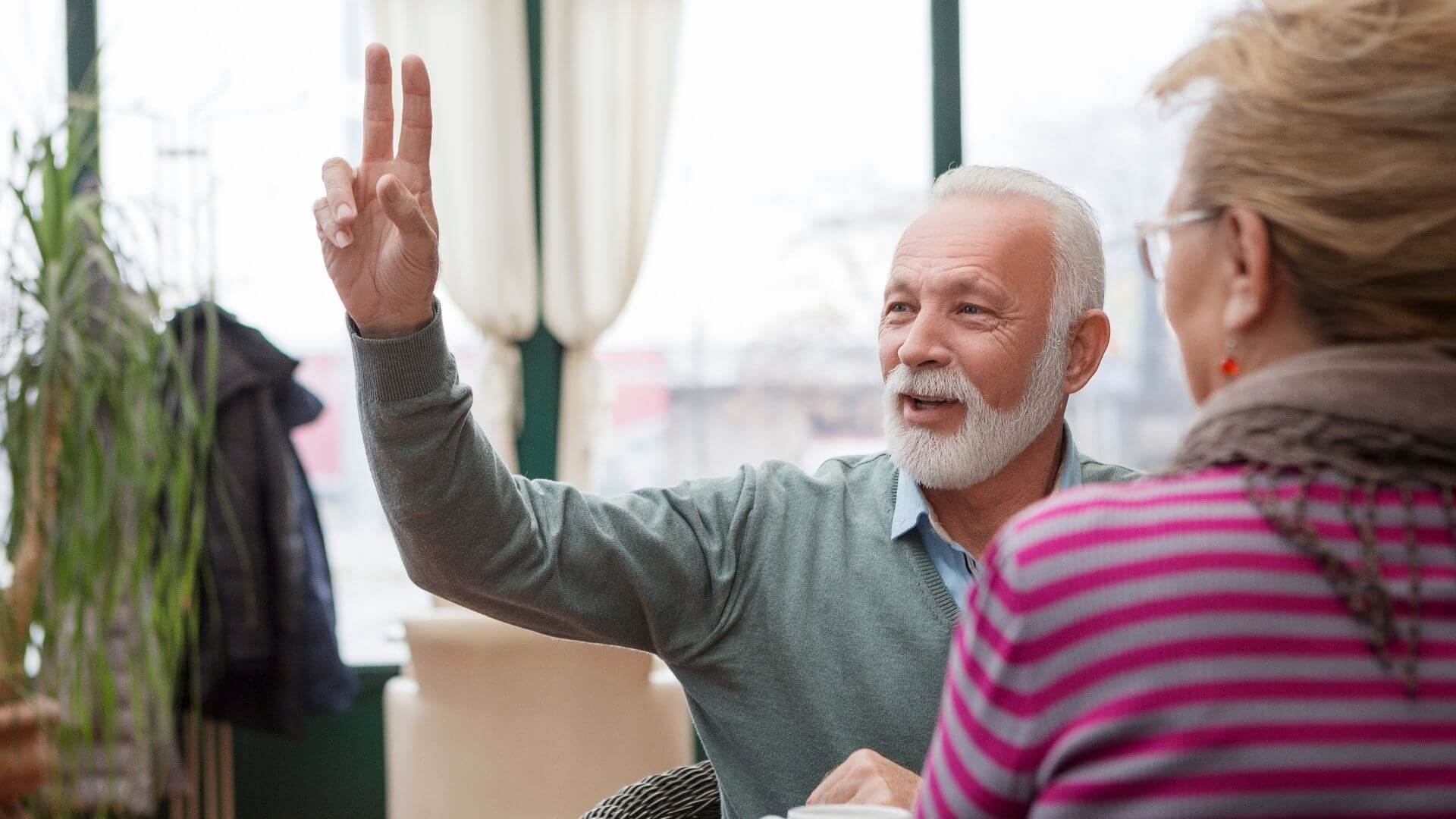 Ein älterer Mann unterhält sich mit einer Frau in einem Café und zeigt mit den Fingern die Zahl zwei.