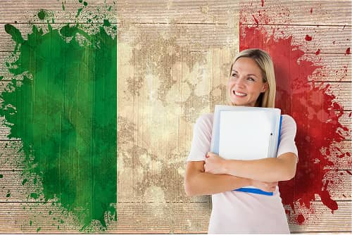 Blonde Studentin mit Büchern lächelt vor gemalter italienischer Flagge auf Holzuntergrund
