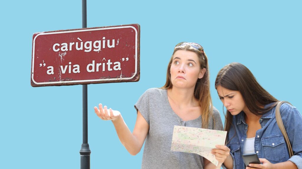 Zwei Frauen stehen neben einem Straßenschild mit der Aufschrift "carùggiu 'a via drîta'"; die eine schaut verwirrt, während die andere eine Karte betrachtet.