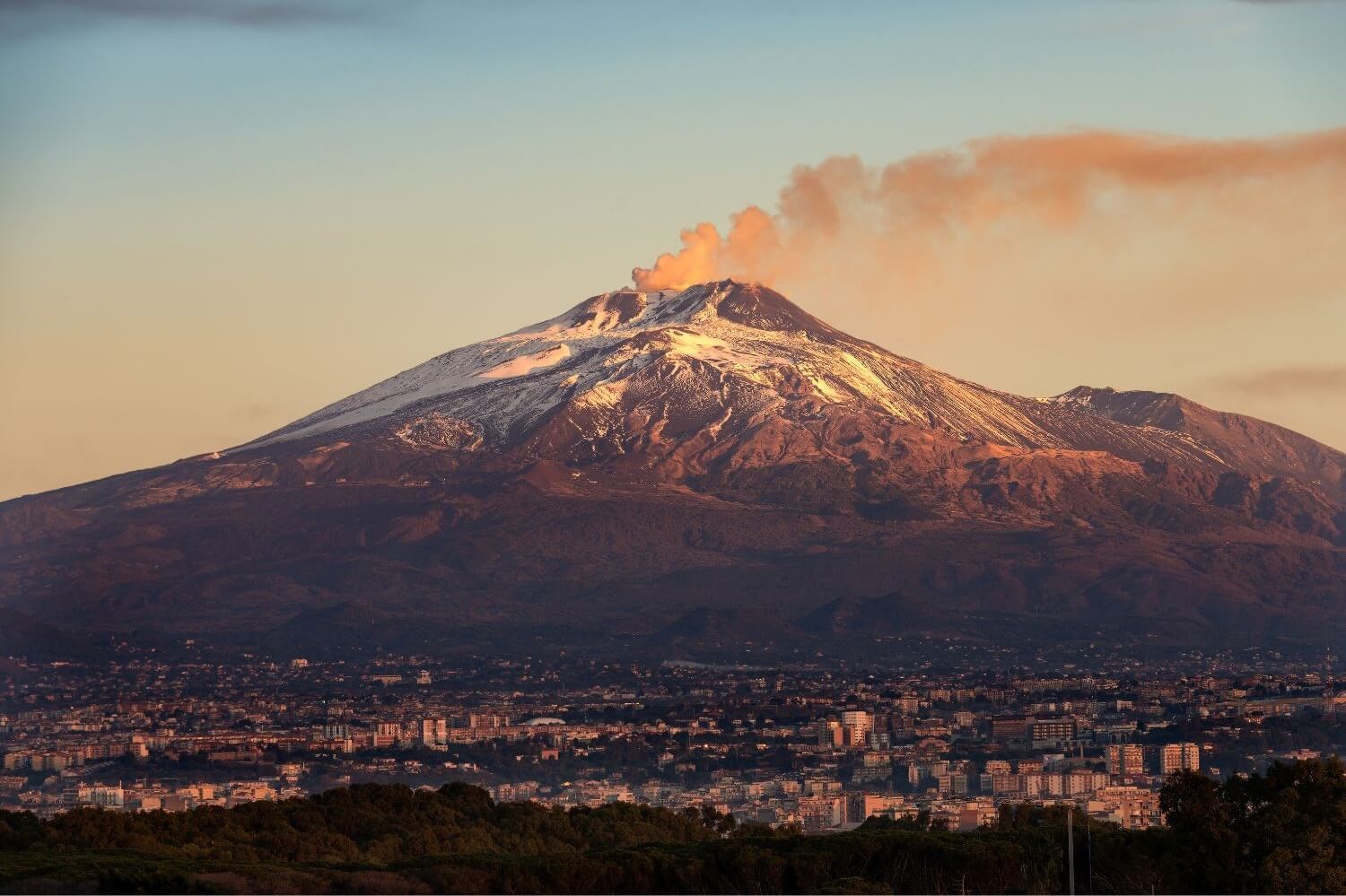 Blick auf den rauchenden Vulkan Ätna bei Sonnenuntergang, mit der Stadt Catania im Vordergrund.