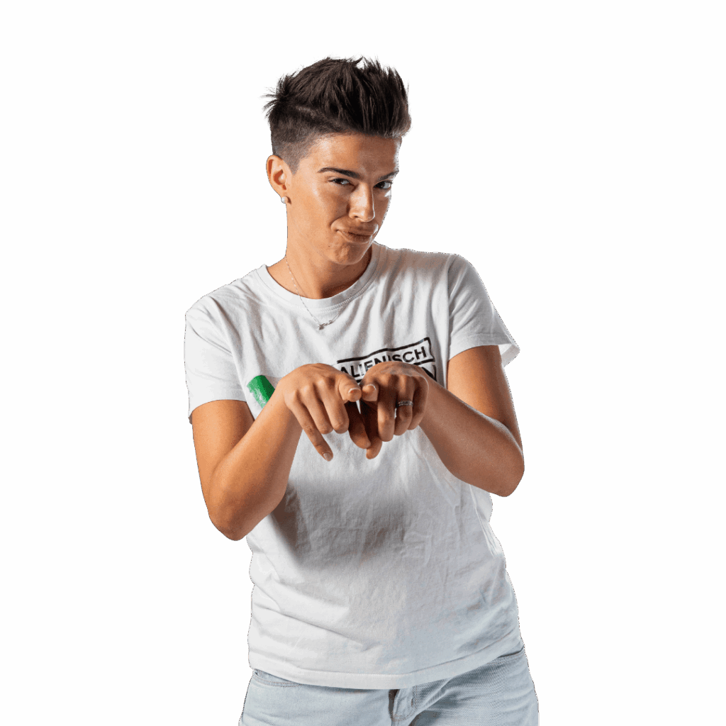 Person mit kurzen dunklen Haaren, die ein weißes T-Shirt und eine helle Hose trägt, steht mit angewinkelten Ellbogen und gefalteten Händen in der klassischen "Se l'intendono"-Geste und macht einen spielerischen oder spöttischen Gesichtsausdruck.