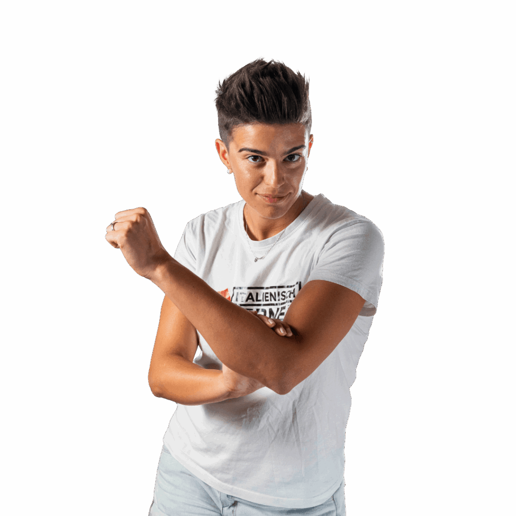 Eine Person mit kurzen dunklen Haaren, die ein weißes T-Shirt und helle Shorts trägt, beugt ihren rechten Arm und macht die "Vaffanculo"-Geste, während sie direkt in die Kamera schaut, vor einem einfarbigen Hintergrund.