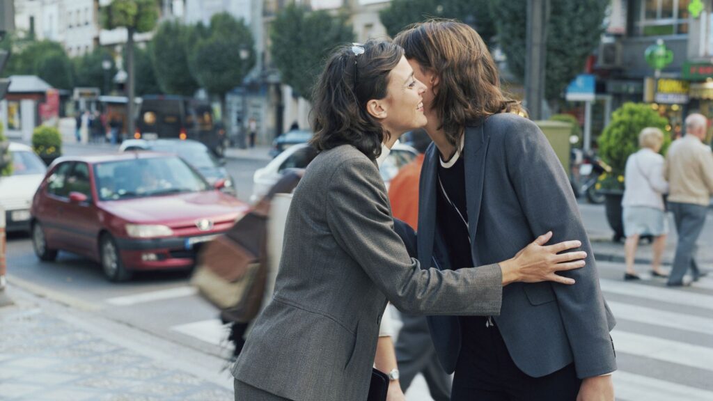 Zwei Frauen in formellen Anzügen begrüßen sich auf der Straße mit einem Kuss auf die Wange.