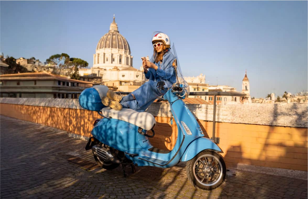 Frau mit Helm sitzt entspannt auf blauer Vespa, im Hintergrund große Domkuppel.