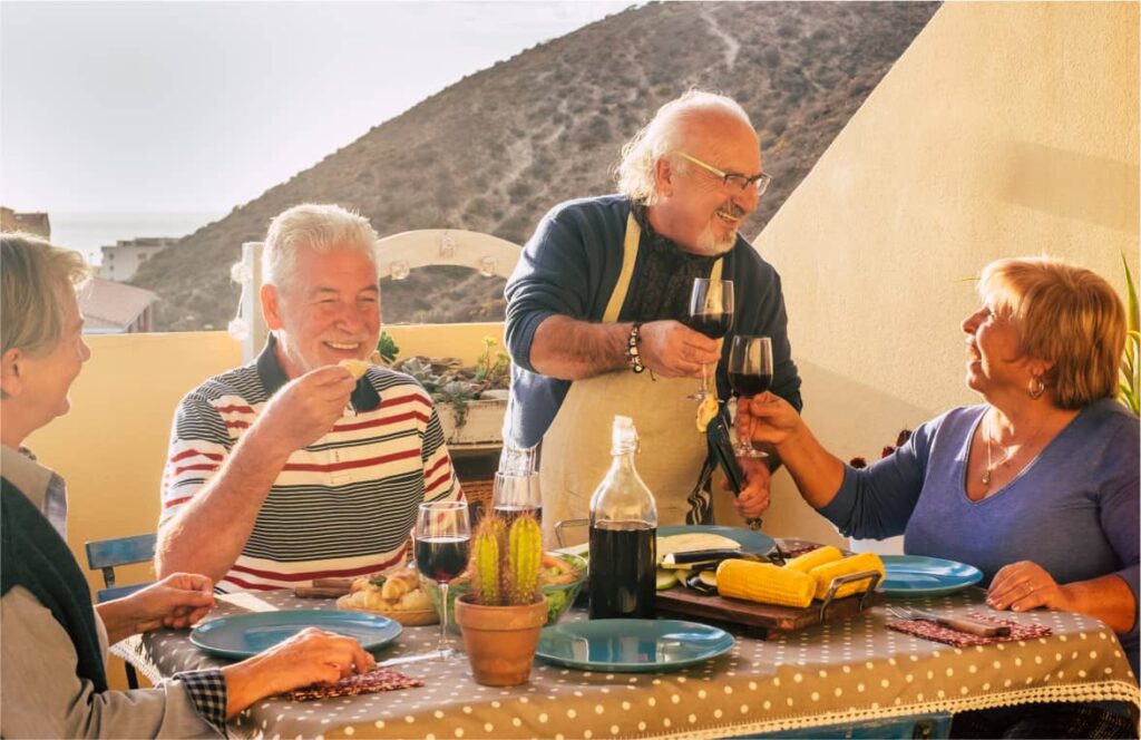 Ältere Menschen sitzen zusammen draußen und stoßen mit Rotwein auf der Terrasse an.