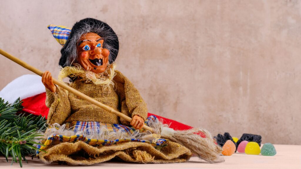 Eine Befana-Hexenpuppe mit einem Besenstiel sitzt auf einem Tisch neben einer Weihnachtsmannmütze, immergrünen Zweigen und bunten Gummibonbons.