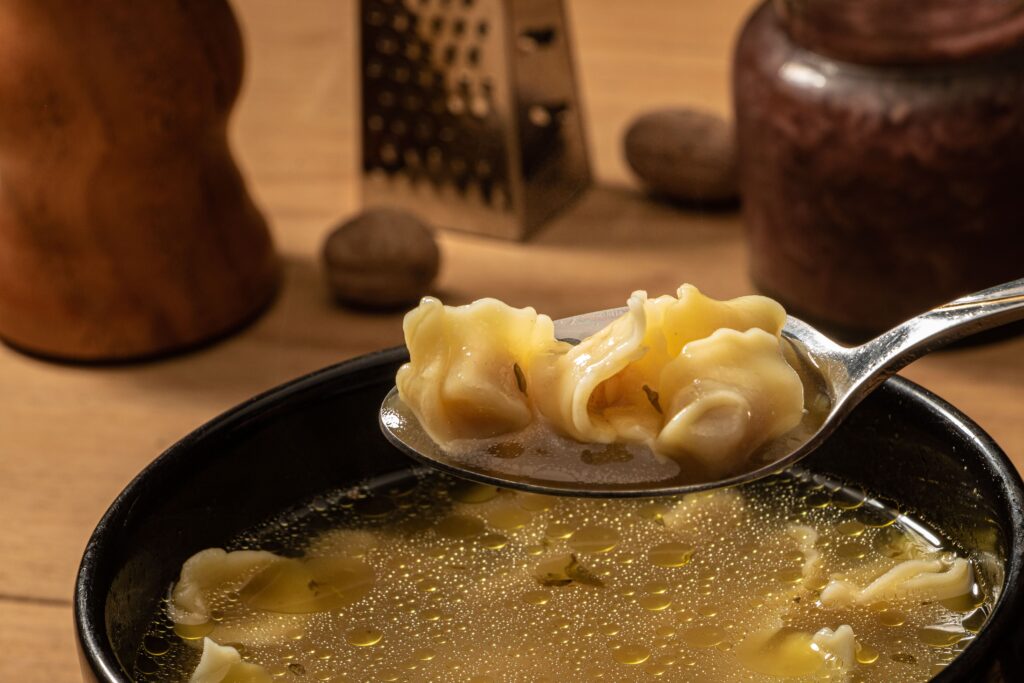 Die Nahaufnahme eines Löffels, der Cappelletti in Brodo über einer Schüssel mit klarer Suppe hält, mit Küchenutensilien, die im Hintergrund verschwimmen, erinnert an die Wärme einer italienischen Weihnachtsfeier.