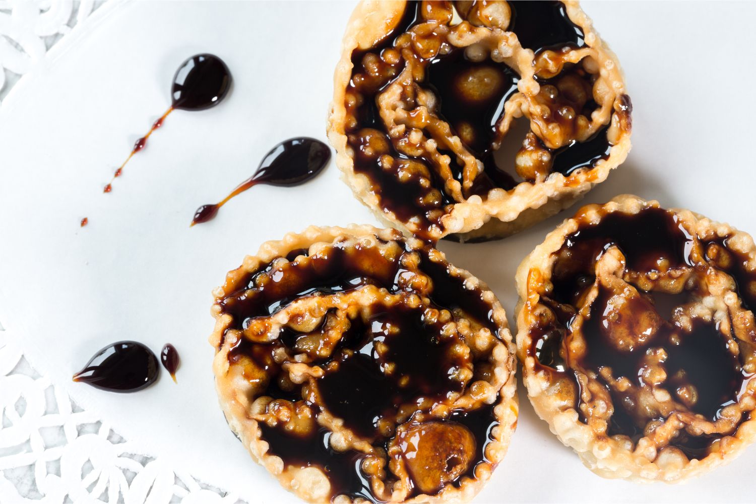 Drei italienische Cartellate Stücke mit dunklem Zuckersirup liegen auf einem weißen Teller mit Siruptropfen und einer mit Spitzen gemusterten Matte.