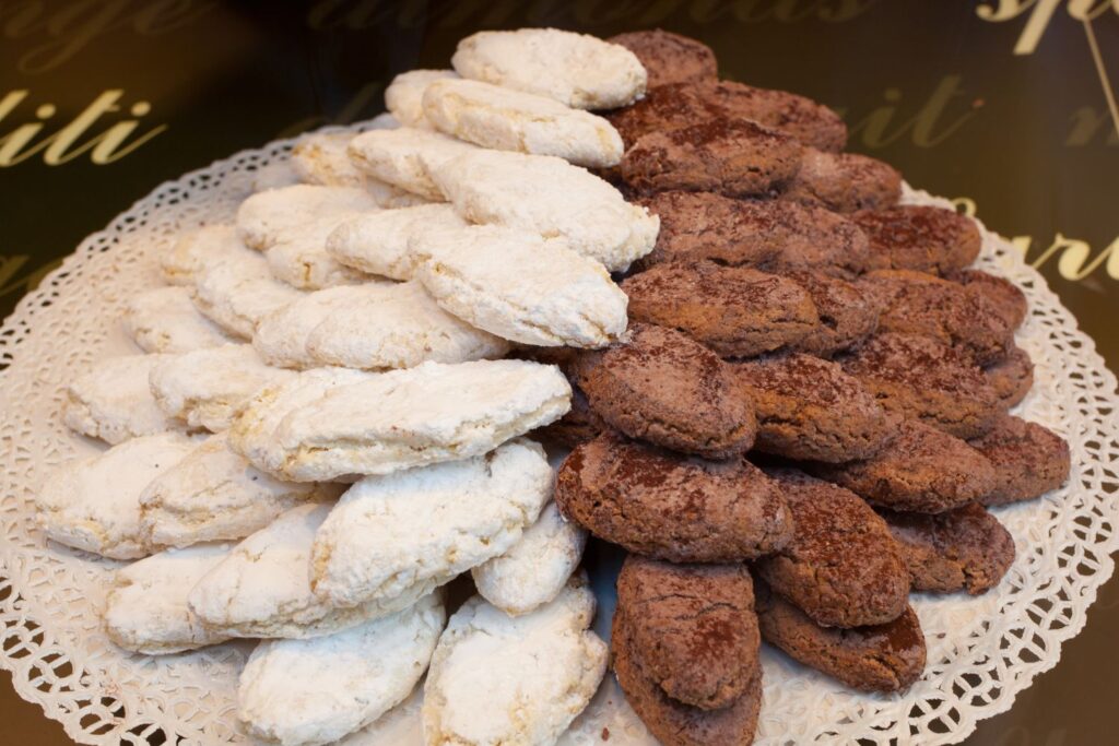 Ein Teller mit ovalen Ricciarelli-Plätzchen, die zur Hälfte mit Puderzucker und zur Hälfte mit Schokolade bestäubt sind und in abwechselnden Reihen auf einem Spitzendeckchen angeordnet sind.