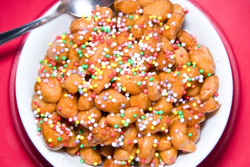 Eine weiße Schale, gefüllt mit Struffoli - mit Honig überzogene, frittierte Teigstücke, die mit bunten, runden Streuseln bestreut sind - steht neben einem Löffel auf einer roten Fläche.