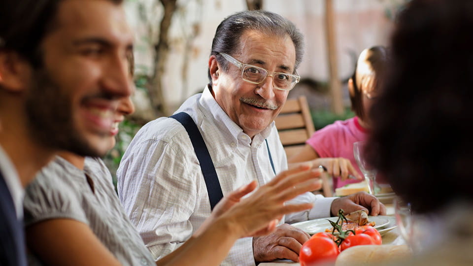 Ein älterer Mann mit Brille und Hosenträgern sitzt lächelnd an einem Tisch im Freien, umgeben von anderen Menschen und Speisen, darunter Tomaten im Vordergrund.