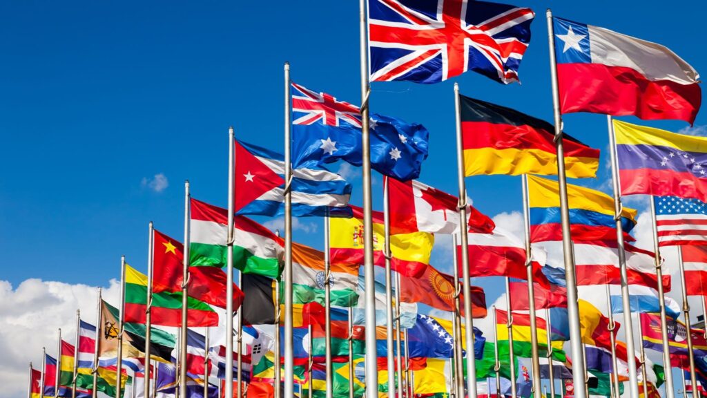 Viele bunte Nationalflaggen aus verschiedenen Ländern wehen auf Fahnenmasten vor einem blauen Himmel.