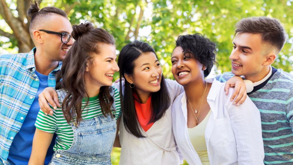 Eine Gruppe junger internationaler Freunde steht lachend zusammen im Park und legt sich die Arme um die Schultern.