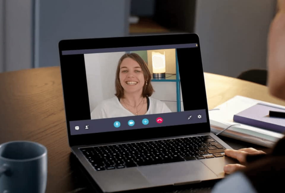 Eine Person, die an einem Videogespräch über einen Laptop teilnimmt, zeigt eine lächelnde Frau auf dem Bildschirm. Auf dem Tisch neben dem Computer stehen eine Tasse und Notizbücher.