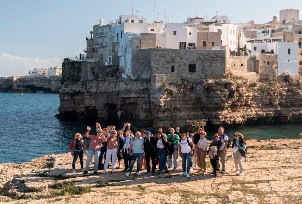 Eine Gruppe von Menschen steht an einem felsigen Ufer in Apulien und winkt in die Kamera, mit weißen Gebäuden auf Klippen und dem Meer im Hintergrund - ein perfekter Moment ihrer Sprachreise Apulien.