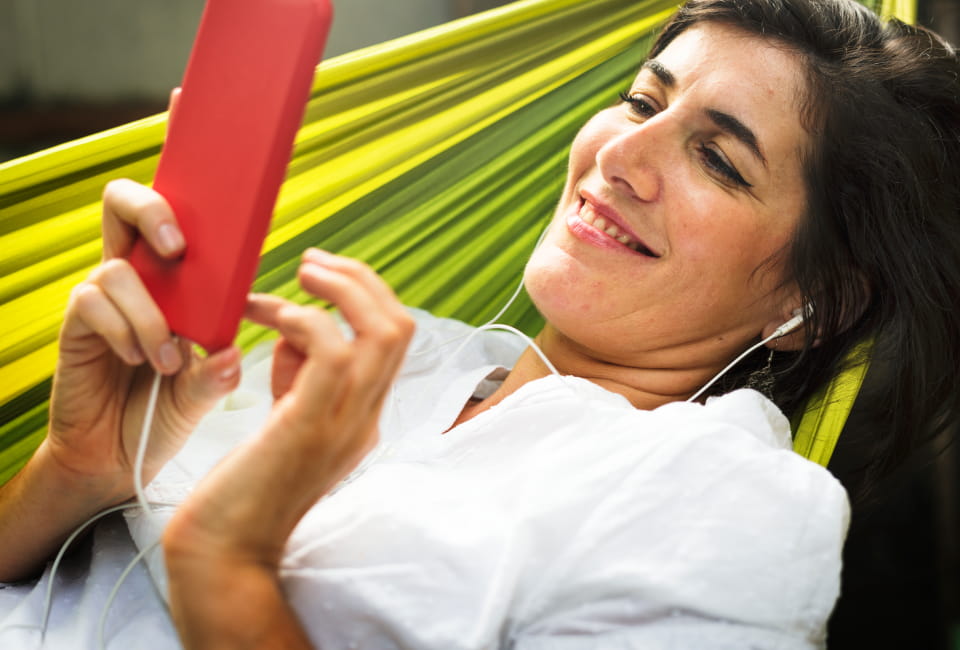 Eine Frau liegt in einer grünen Hängematte, trägt Kopfhörer und lächelt, während sie einen italienischen Audio-Newsletter auf ihrem roten Telefon anhört.