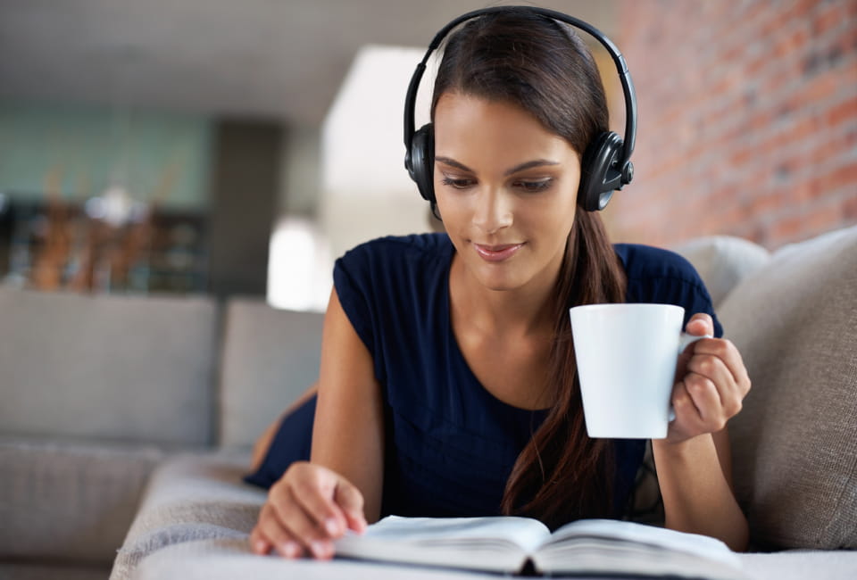 Eine Frau mit Kopfhörern liegt auf einer Couch, genießt einen italienischen Audio-Newsletter, hält eine weiße Tasse in der einen Hand und liest mit der anderen ein aufgeschlagenes Buch.