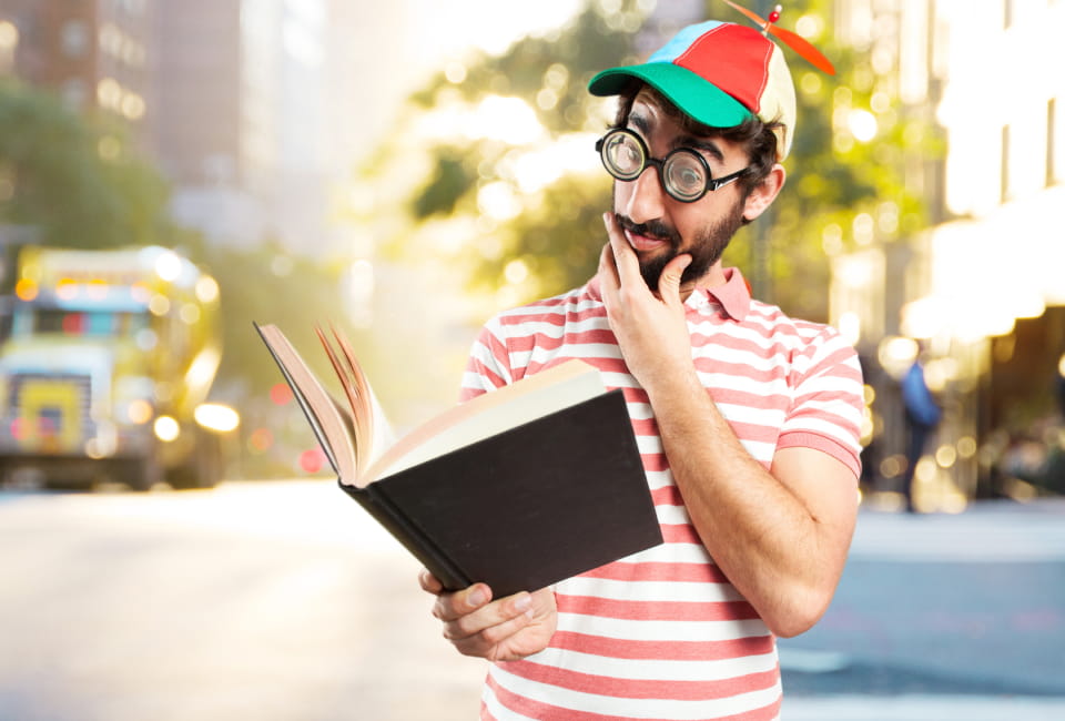 Ein Mann mit einem bunten Propellerhut und einer Scherzbrille steht im Freien und liest mit einem verwirrten Gesichtsausdruck ein Buch. Im Hintergrund ist ein Schulbus zu sehen.