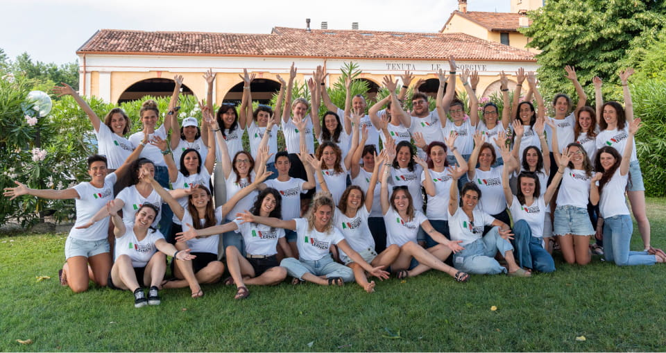 Das Team von italienisch lernen mit Francesca in passenden weißen Hemden posiert im Freien mit erhobenen Armen, sitzend und stehend im Gras vor einem begrünten Gebäude.
