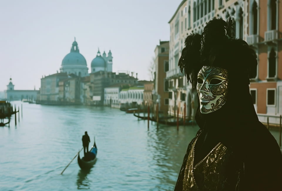 Eine Person mit einer verzierten venezianischen Maske und Kostüm steht an einem Kanal in Venedig, mit einer Gondel und historischen Gebäuden im Hintergrund.